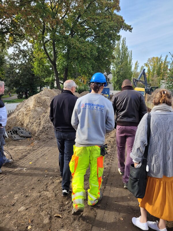 Mehrere Personen gehen gemeinsam über eine Baustelle. Im Vordergrund trägt ein Mitarbeiter von Frisch & Faust Tiefbau einen blauen Schutzhelm und auffällige Warnkleidung; im Hintergrund sind Bagger, Erdhaufen und Baustelleneinrichtungen zu sehen.