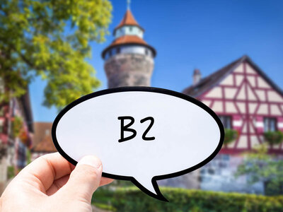Eine Hand hält eine Sprechblase mit der Aufschrift 'B2' in den Vordergrund. Im Hintergrund ist ein historisches Fachwerkhaus mit einem Turm zu sehen. Das Haus hat ein traditionelles Design mit roten Balken und weißem Verputz. Der Turm hat ein spitzes Dach und ist von grünen Bäumen und einem klaren blauen Himmel umgeben.
