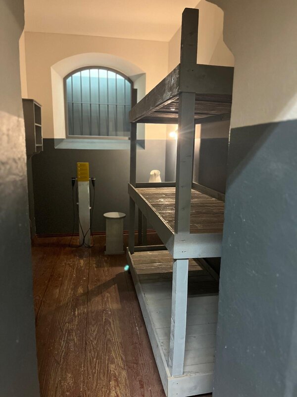 Blick in eine kleine, spärlich eingerichtete Zelle mit Holz-Doppelstockbett, vergittertem Fenster und einfacher Ausstattung. Der Raum wirkt authentisch erhalten oder rekonstruiert.