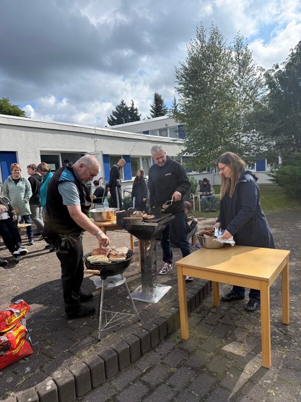 Im Vordergrund stehen zwei Männer und eine Frau an einem Grill und einem Holztisch. Die Männer grillen Würstchen und Fleisch, während die Frau am Tisch eine Packung öffnet. Im Hintergrund sind mehrere Menschen zu sehen, die sich auf einem gepflasterten Platz vor einem weißen Gebäude mit blauen Fensterrahmen aufhalten. Der Himmel ist bewölkt, und einige Bäume rahmen die Szene ein.