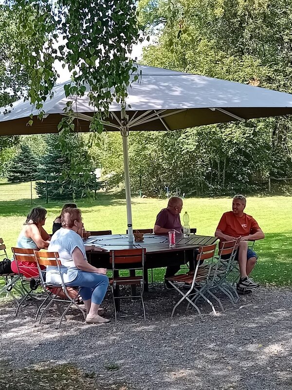 Unter einem großen Sonnenschirm im Schatten am Rand einer Wiese sitzen an einem großen runden Tisch drei Frauen und zwei Männer. Auf dem Tisch stehen ein Glas und eine Plastikflasche.