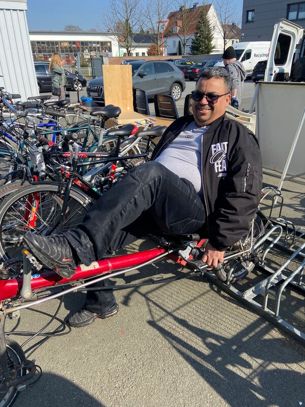 Ein lächelnder Mann sitzt draußen zwischen zahlreichen Fahrrädern auf einem roten Liegefahrrad oder Tandem und probiert es aus.