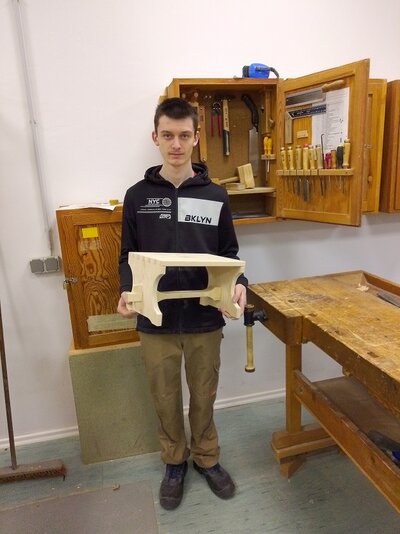 Ein junger Mann steht neben einer Werkbank und hält seine selbstgebaute Fußbank aus Holz in den Händen