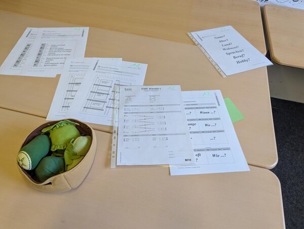 Auf einem hellen Tisch liegen mehrere ausgedruckte Arbeitsblätter, teils mit Tabellen und englischsprachigen Fragen. Links steht ein kleiner Korb mit mehreren grünen und blauen Stoffbällen. In der rechten oberen Ecke ist ein weiteres Blatt mit großformatigem Text zu sehen.