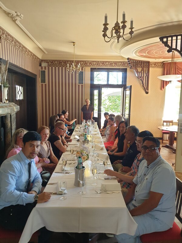 Gruppe von Erwachsenen sitzt an einer langen, festlich gedeckten Tafel in einem historischen Restaurant und blickt teilweise in die Kamera.