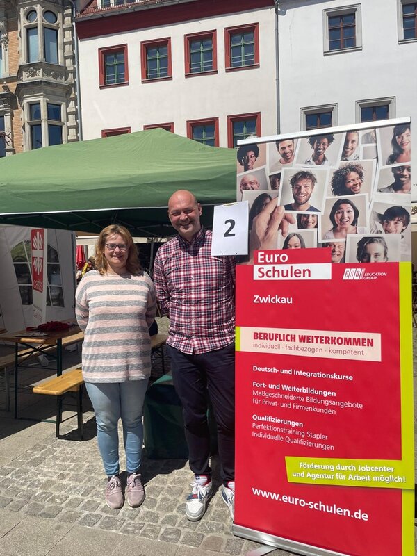 Mitarbeitende der Euro-Schulen Zwickau posieren lächelnd für ein gemeinsames Bild vor ihrem Euro-Schulen Infostand auf dem Interkulturellen Familienfest in Zwickau. 