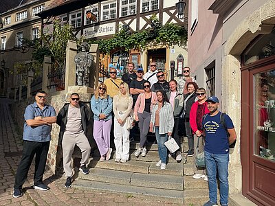 Ausflug in Meißen: eine Märchenstadt an der Elbe Eine Gruppe von Menschen posiert auf einer Steintreppe vor einem historischen Fachwerkhaus mit dem Schriftzug „Vinzenz Richter“. Neben der Gruppe steht eine Statue, und die Szene ist von Sonnenschein erhellt.