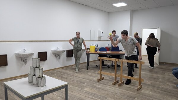 Dosenwerfen in einem Klassenraum. Ein junger Mann in grauem Pullover wirft einen Tennisball auf die Dosen.