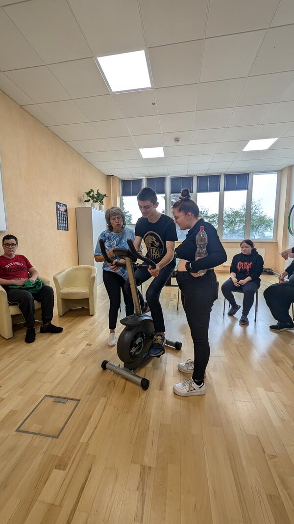 Eine Gruppe von Schülerinnen und Schülern stehen zusammen mit einer Lehrkraft um einen Heimtrainer. Darauf sitzt ein Schüler, der interessiert auf den Bildschirm des Heimtrainers schaut.