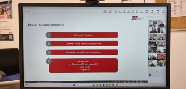 Ein Computerbildschirm zeigt eine Präsentationsfolie mit dem Titel 'OKLUMA – BESONDERHEITEN FÜR VK'. Die Folie enthält vier nummerierte Punkte in grauen und roten Feldern: 'Start- und Enddatum', 'Lehrkraft in die Veranstaltung buchen', 'Teilnehmer: Mailadressen hinterlegen' und 'Stundenplan: Zeitraster (Block-Unterricht), Jitsi-Meet, Freigabe VC'. Oben rechts sind mehrere kleine Videofenster mit Personen in einer Videokonferenz zu sehen. Im Hintergrund sind ein Teil eines Schreibtisches, ein Stuhl und eine Pinnwand mit verschiedenen Zetteln erkennbar.