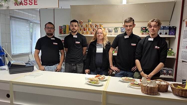 Alles neu macht der… September Vier Schüler in Berufsschutzbekleidung stehen mit ihrer Lehrerin hinter der Verkaufstheke. Auf der Theke stehen verschiedene belegte Brötchen, in dem Regal hinter den Personen weitere abgepackte Lebensmittel. Links steht eine Kasse.
