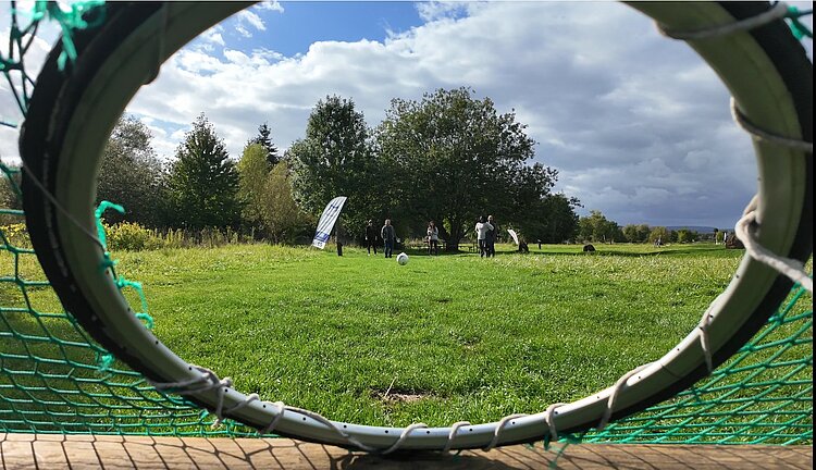 Team-Building beim Fußballgolf Andere Blickwinkel: Durch ein Fußballnetz, in den ein Ring integriert ist sieht man die Teilnehmenden auf der Wiese vor ein paar Bäumen stehen. Ein Fußball fliegt in Richtung des Ringes, auf die Kamera zu.