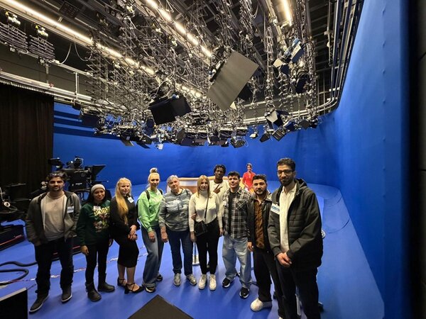 Die  Gruppe der Teilnehmenden des Integrationskurses steht in einem Fernsehstudio mit komplett blauen Wänden und Boden. Über ihnen hängen zahlreiche Scheinwerfer und technische Geräte an einer komplexen Deckenkonstruktion. Im Hintergrund sind Kameras und weiteres Studioequipment sichtbar. Die Personen stehen nebeneinander und blicken in Richtung Kamera.