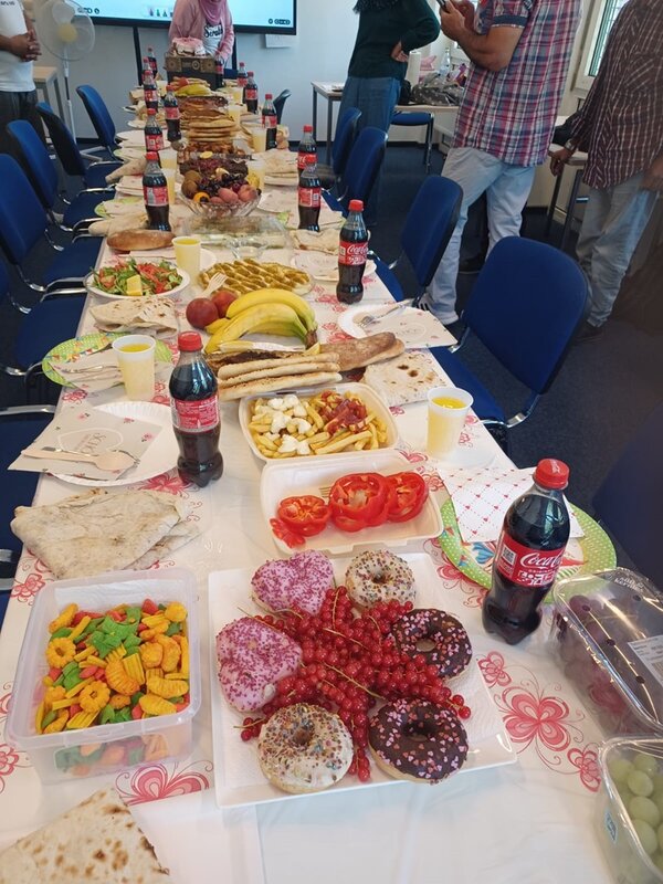 Lange gedeckte Tafel in einem Seminarraum mit zahlreichen Speisen, darunter Fladenbrot, Salate, Obst, Pommes, Gemüse, Donuts und weitere Backwaren sowie Getränkeflaschen.
