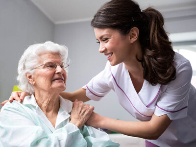 Zwei Personen interagieren miteinander. Eine Person scheint eine ältere Person zu sein, die sitzt, während die andere Person, die wie eine Pflegekraft oder ein medizinischer Fachmann aussieht, steht und sanft die Hände auf die Schultern der älteren Person legt. Die Gesichter beider Personen sind aus Datenschutzgründen unscharf. Die Pflegekraft trägt eine weiße Uniform mit lila Besatz.