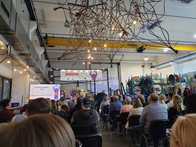 Blick in eine große Industriehalle während der Verleihung des Reinickendorfer Ausbildungsbuddy-Preises. Zahlreiche Gäste sitzen vor einer Bühne, über der ein großes Kletternetz und ein gelber Hallenkran hängen.