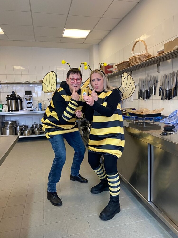 Zwei Mitarbeitende in Bienenkostümen posieren fröhlich in einer modernen Küche mit einem Glas Honig in der Hand für ein Foto.