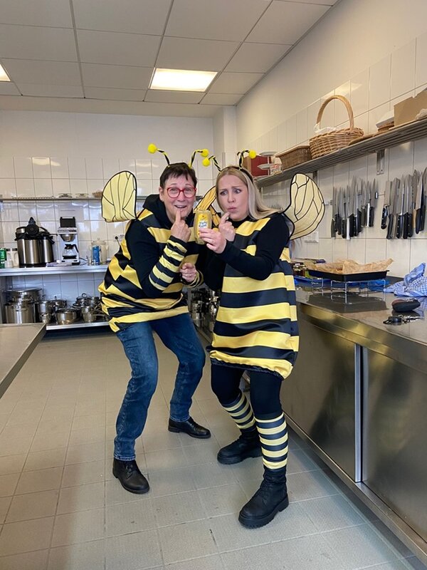 Zwei Mitarbeitende in Bienenkostümen posieren fröhlich in einer modernen Küche mit einem Glas Honig in der Hand für ein Foto.