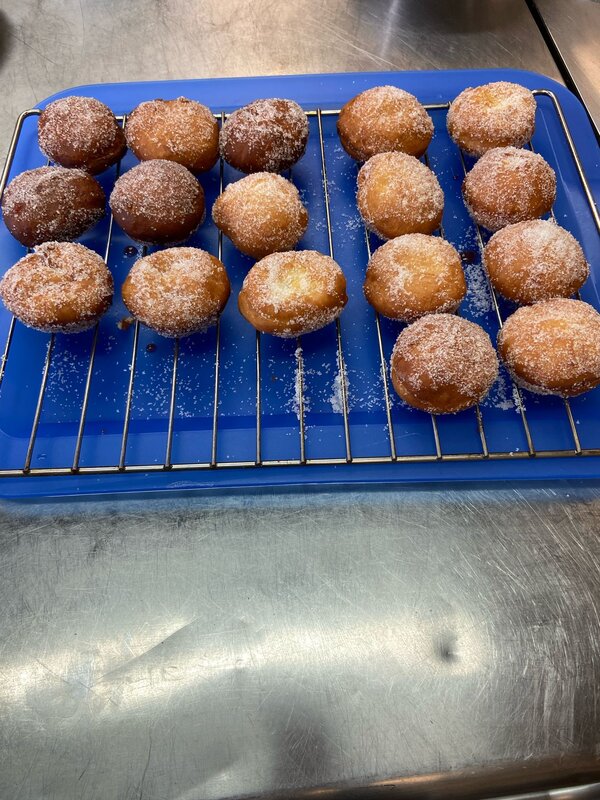 Frisch gebackene, mit Zucker bestreute Krapfen liegen auf einem blauen Gitter und kühlen aus. Einige sind dunkler gebacken als andere, und auf der Arbeitsfläche befinden sich Zuckerkristalle.