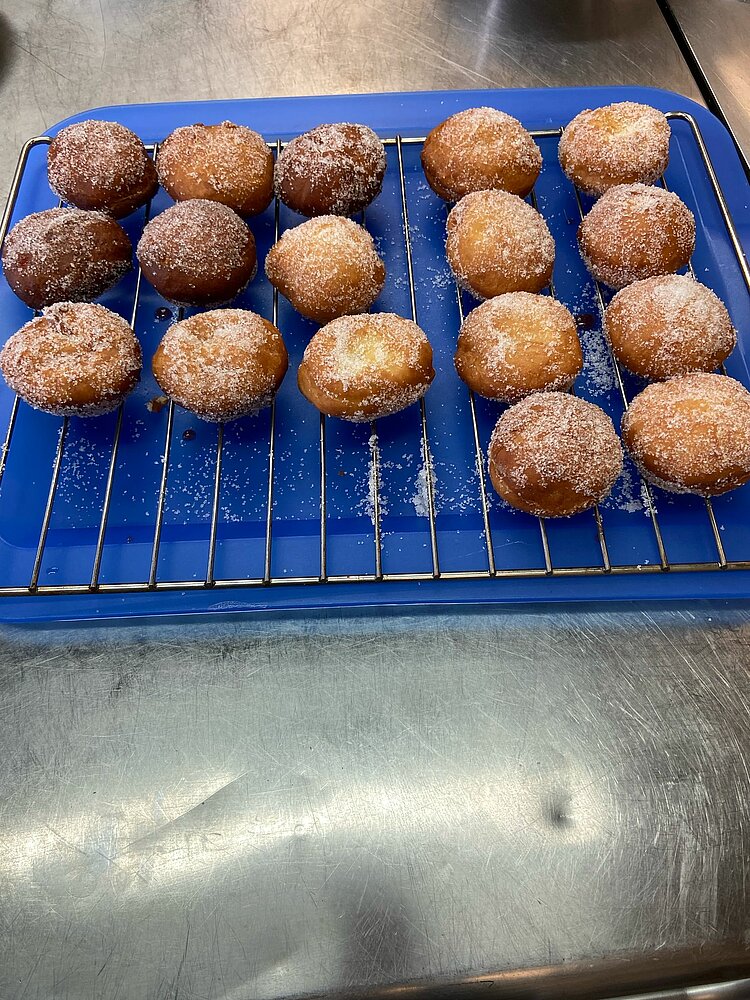 Kein Karneval in Bielefeld Frisch gebackene, mit Zucker bestreute Krapfen liegen auf einem blauen Gitter und kühlen aus. Einige sind dunkler gebacken als andere, und auf der Arbeitsfläche befinden sich Zuckerkristalle.