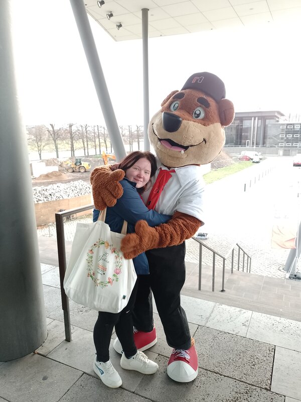 Nahaufnahme: Teilnehmerin umarmt lächelnd das Bären-Maskottchen auf der Treppe; modernes Gebäude und Außengelände im Hintergrund.