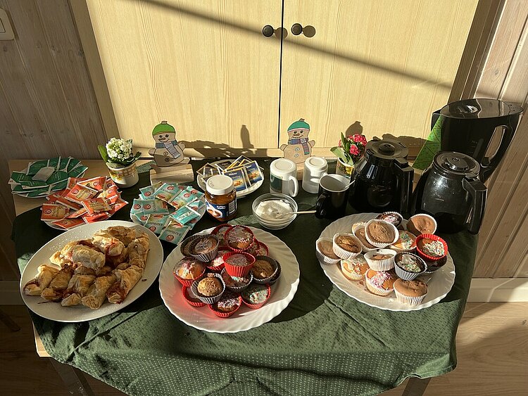 Sprachcafé und Tee – Ein Ort des Austauschs und der Verständigung Auf einem Tisch stehen drei großes Teller mit unterschiedlichem Gebäck (u.a. Muffins) sowie mehrere Teller mit unterschiedlichen Teesorten und Zucker sowie Kannen und ein Wasserkocher. Zur Deko stehen kleine Blümchen und Schneemann-Figuren auf dem Tisch.