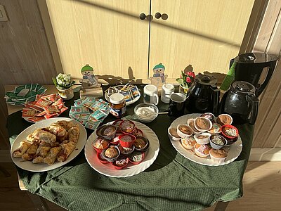 Sprachcafé und Tee – Ein Ort des Austauschs und der Verständigung Auf einem Tisch stehen drei großes Teller mit unterschiedlichem Gebäck (u.a. Muffins) sowie mehrere Teller mit unterschiedlichen Teesorten und Zucker sowie Kannen und ein Wasserkocher. Zur Deko stehen kleine Blümchen und Schneemann-Figuren auf dem Tisch.