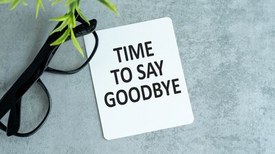 Auf einer grauen Oberfläche liegt eine weiße Karte mit dem gut lesbaren, schwarzen Text 'TIME TO SAY GOODBYE'. Links neben der Karte befindet sich eine schwarze Brille, daneben ragt ein grüner Pflanzenzweig ins Bild. Die Szene vermittelt einen Abschiedsmoment, passend zur Verabschiedung der langjährigen Schulleiterin der Euro-Schulen Aschaffenburg.