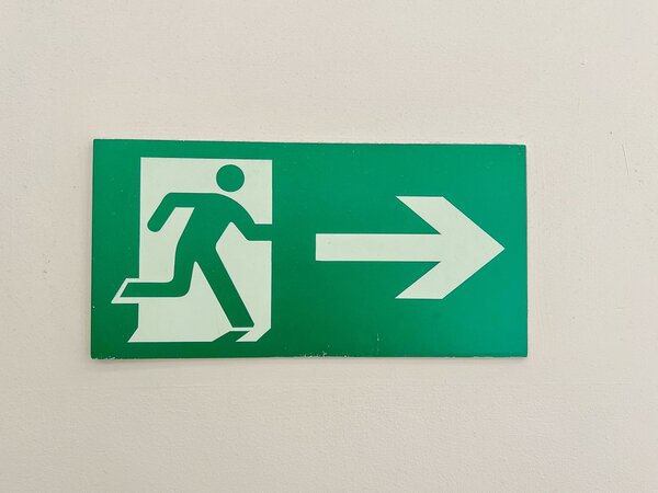 Grünes Notausgang-Schild mit weißem Piktogramm einer laufenden Person, die durch eine Tür nach rechts geht. Rechts neben der Figur befindet sich ein großer, weißer Pfeil, der nach rechts zeigt. Das Schild ist auf einer hellen Wand angebracht.