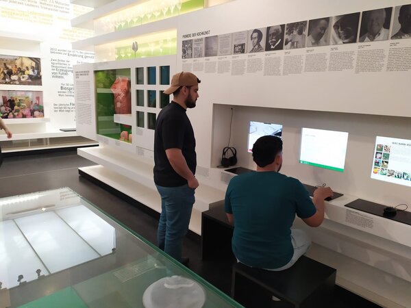 Zwei Personen in einem modernen Ausstellungsraum sitzen an Computern und lesen oder recherchieren Informationen zu einem Thema.