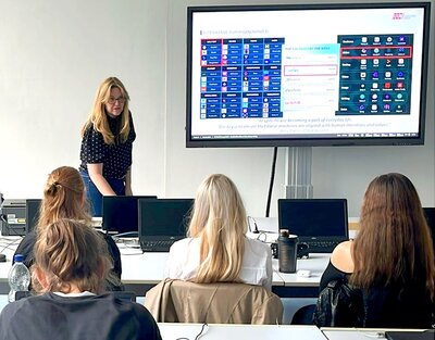 Workshopleiterin steht vorn im Klassenraum und referiert vor Schülerinnen und Schülern über Künstliche Intelligenz. 