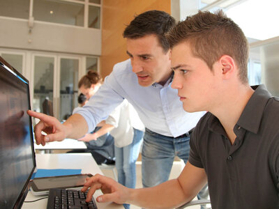 Zwei Personen, ein Mann und ein Jugendlicher, betrachten gemeinsam einen Computerbildschirm. Der Mann zeigt auf den Bildschirm, während der Jugendliche die Tastatur bedient. Im Hintergrund ist eine weitere Person an einem Schreibtisch zu sehen.