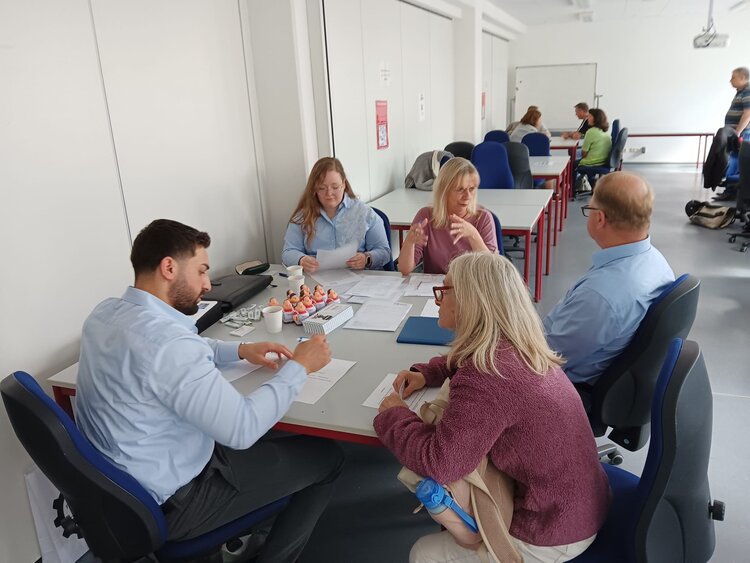 Eine Kleingruppe sitzt an einem Tisch in einem Seminarraum und bespricht Unterlagen. Auf dem Tisch befinden sich Papiere, Schreibmaterialien und kleine Figuren. Eine Frau erklärt etwas mit Gesten, während die anderen aufmerksam zuhören.