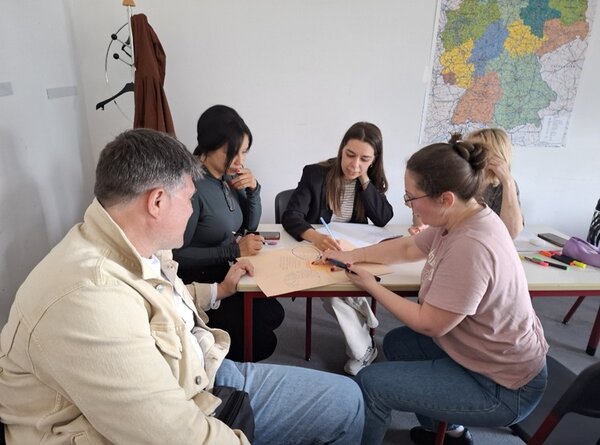 Weibliche und männliche Teilnehmende des B2-Kurses sitzen an einem großen Tisch und arbeiten an ihren zu erstellenden Aufgaben.