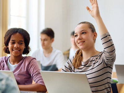 Das Bild zeigt eine Unterrichtssituation. Im Vordergrund sitzen zwei Mädchen vor aufgeklappten Laptops an ihren Tischen. Das Mädchen links ist dunkelhäutig und trägt einen rosefarbenen Pullover. Das Mädchen rechts mit gestreiftem T-Shirt hebt die linke Hand und schaut erwartungsvoll nach vorn