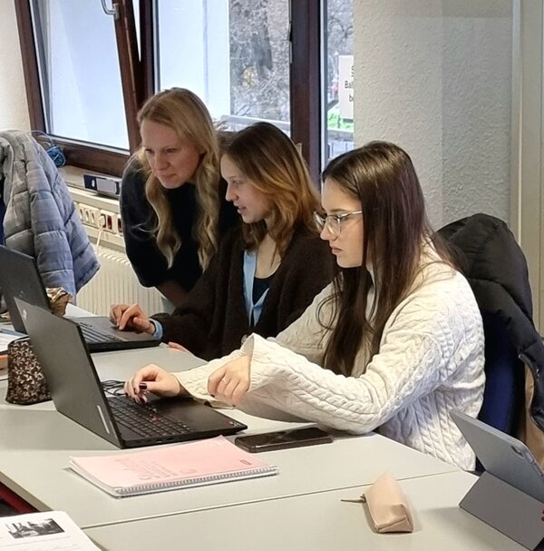 Zwei Personen sitzen zusammen an einem Tisch und arbeiten konzentriert an Laptops. Eine Person steht hinter ihnen und schaut auf die Bildschirme, möglicherweise um Unterstützung oder Anleitung zu geben. Der Raum ist hell und hat große Fenster, durch die Tageslicht einfällt. Auf dem Tisch liegen Notizblöcke, Stifte und persönliche Gegenstände wie ein Handy und eine Tasse.