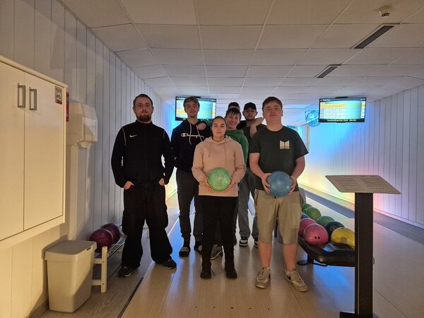 Gruppenfoto auf der Bowlingbahn: Mehrere Teilnehmende stehen gemeinsam am Bahnanfang, zwei halten Bowlingkugeln in den Händen; im Hintergrund sind Anzeigetafeln und Kugelrücklauf zu sehen.