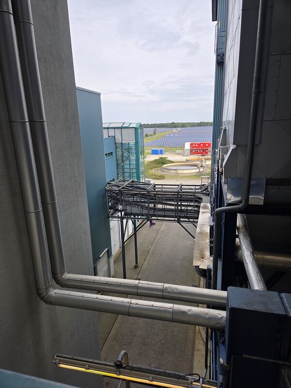 Blick von einer erhöhten Position zwischen Industriegebäuden auf Rohrleitungen, einen Metallsteg und das Betriebsgelände. Im Hintergrund sind ein großes Feld mit Solarmodulen, ein rotes Gebäude und runde technische Becken zu sehen.