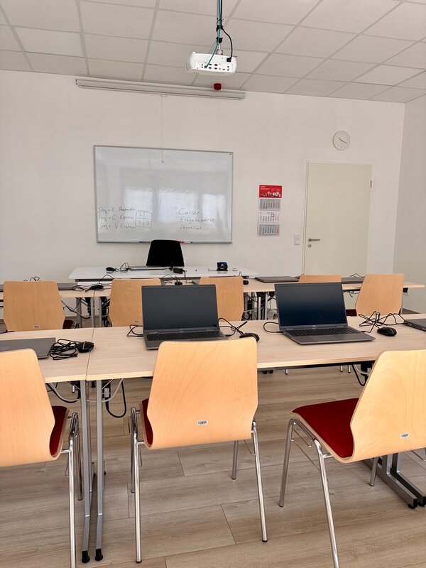 Moderner Unterrichtsraum mit Laptops, Beamer, Whiteboard und vorbereiteten Arbeitsplätzen für den digitalen Unterricht.