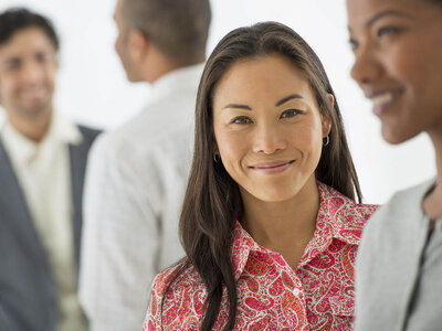 Eine lächelnde Frau mit langen dunklen Haaren und einem gemusterten roten Oberteil steht im Vordergrund. Im Hintergrund sind mehrere Personen in einem Gespräch, die Geschäftskleidung tragen.