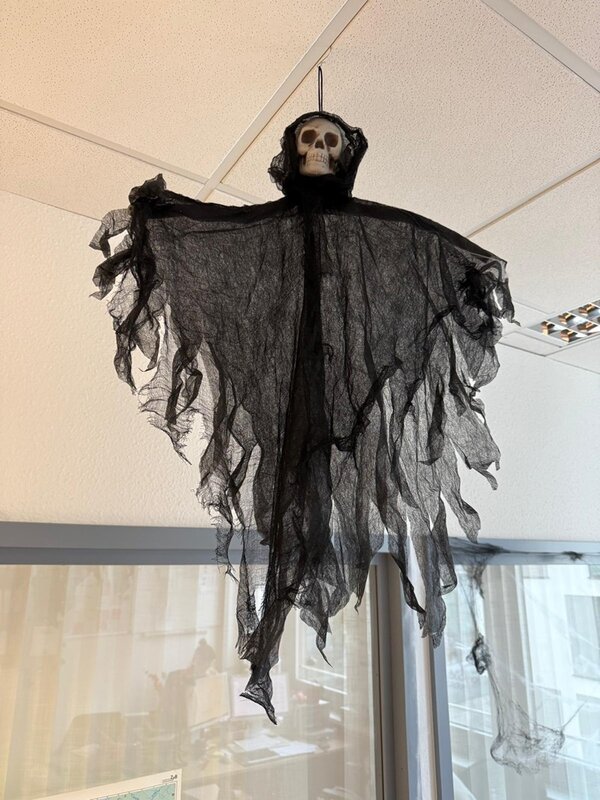 Eine Skelettfigur mit Kapuze und ausgebreiteten Armen hängt von der Decke. Der Körper besteht aus schwarzem, durchscheinendem Stoff, der in Fetzen herabhängt. Der Totenkopf ist hell und hebt sich deutlich vom dunklen Stoff ab. Im Hintergrund sind eine weiße Decke mit Deckenplatten und ein Teil eines Fensters zu sehen.