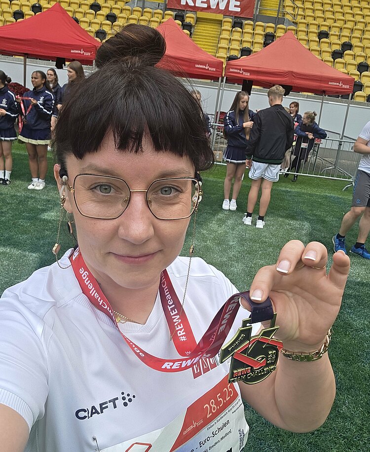 Euro-Schulen & Euro Akademie bei der Rewe Team Challenge 2025 Selfie einer Teilnehmerin, die stolz ihre Finisher-Medaille der REWE Team Challenge zeigt. Im Hintergrund: das Stadion mit roten Zelten und Cheerleadern.