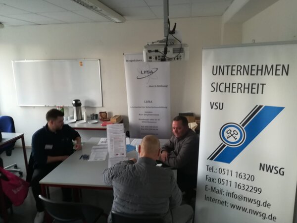 Beratungsgespräch an einem Tisch mit Formularen; daneben Roll-up-Banner „Unternehmen Sicherheit VSU“ und „LiFSA“ im Schulungsraum.