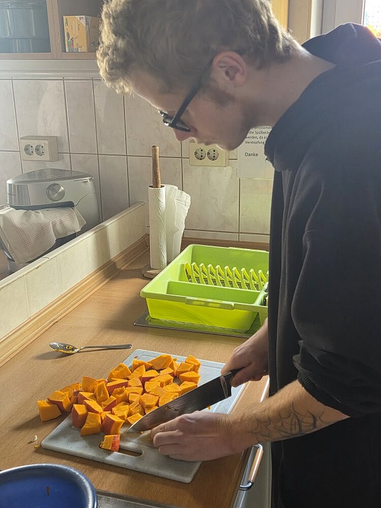 Herbstliche Köstlichkeit Ein Schüler schneidet auf einem Kunststoffbrett mit einem großen Messer den Kürbis in kleine Stücke. Auf der Arbeitsplatte liegt außerdem ein Löffel, mit dem er den Kürbis wohl vorher ausgehöhlt hat.