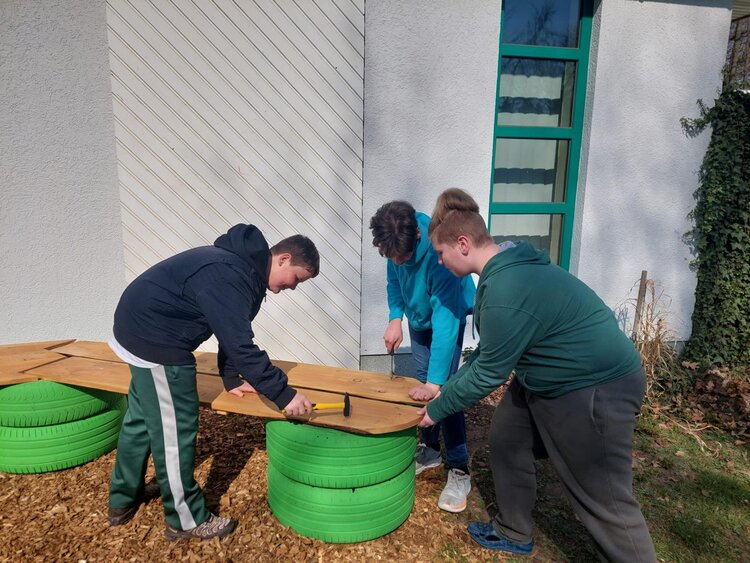 Drei Jugendliche arbeiten gemeinsam an einer Bankkonstruktion aus Holz und grün lackierten Reifen vor einem Gebäude.