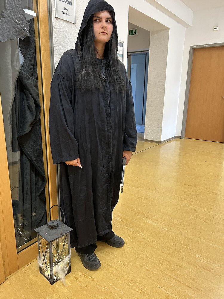 Person in schwarzer, hooded Robe steht im Flur neben einer Laterne mit Lichterkette.