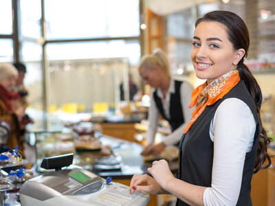 Eine junge Frau mit langen dunklen Haaren und einem orangen Schal steht lächelnd an einer Kasse in einem Café oder einer Bäckerei. Sie trägt ein schwarze Veste über einem weißen Langarm-Shirt. Im Hintergrund sind weitere Mitarbeiter und Kunden zu sehen, die sich an der Theke bedienen. Auf der Theke liegen verschiedene Backwaren und ein Kassenterminal.