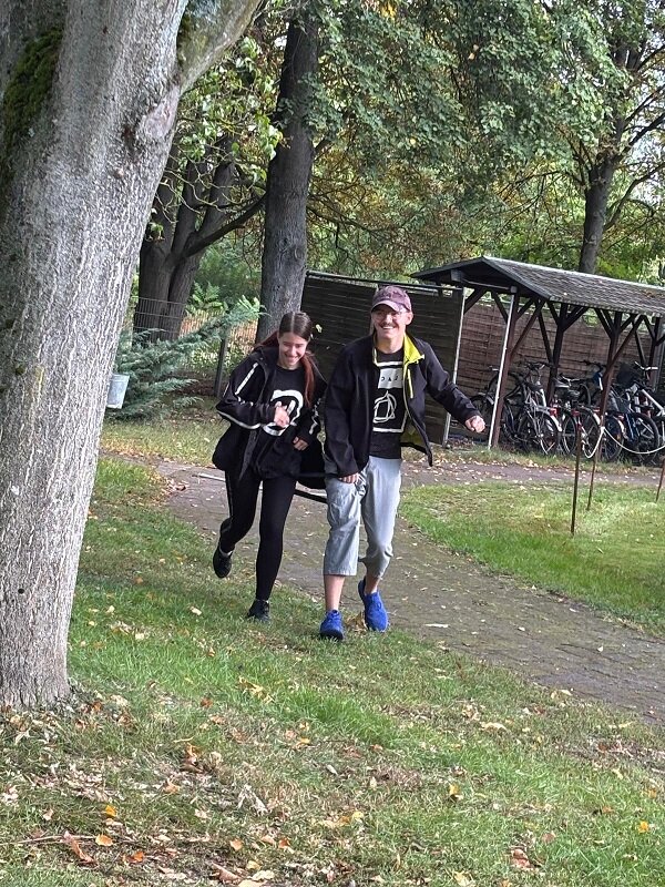 Zwei Personen gehen auf einem schmalen Weg durch eine Grünanlage. Beide tragen dunkle Kleidung und T-Shirts mit auffälligen weißen Motiven. Die vordere Person trägt eine helle Kappe, eine dunkle Jacke, helle Shorts und blaue Schuhe. Die hintere Person trägt eine dunkle Jacke und dunkle Hose. Im Hintergrund stehen Fahrräder unter einem überdachten Fahrradständer, daneben sind Bäume und Rasenflächen zu sehen. Herbstlaub liegt auf dem Boden.