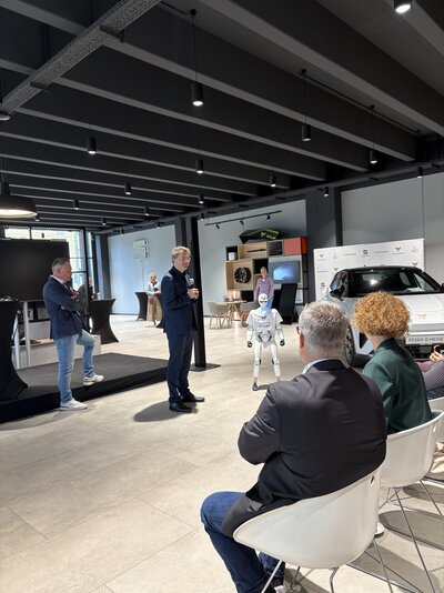 Veranstaltung in einem modernen Autohaus: Ein Mann hält vor sitzenden Gästen einen Vortrag, während neben ihm ein humanoider Roboter steht. Im Hintergrund ist ein weißes Fahrzeug der Marke CUPRA zu sehen.