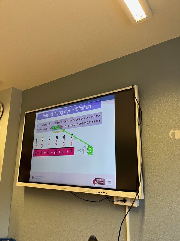 Ein Monitor im Schulungsraum zeigt eine polizeiliche Präsentation zur Berechnung von Prüfziffern in Ausweisdokumenten. Grafiken mit Zahlenreihen, Multiplikationsmustern und Hervorhebungen in Grün und Pink erklären die korrekte Prüfziffer 9. Das Bild dokumentiert die Fortbildung zur Dokumentenprüfung an den Euro-Schulen Hannover.